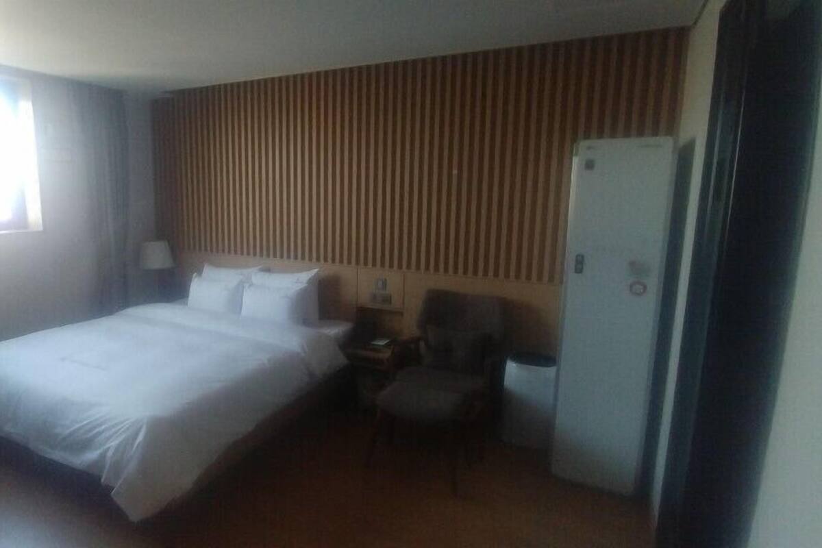 A hotel room with a bed, a chair, a desk, a lamp, and a refrigerator.