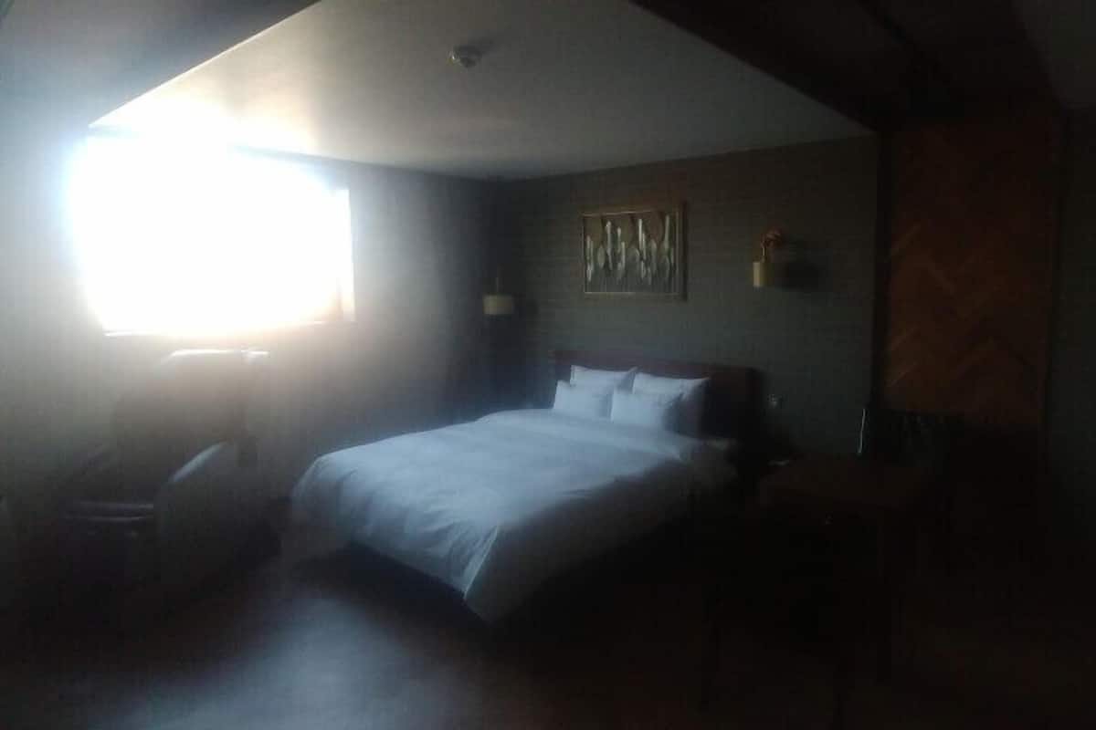 A dimly lit hotel room with a large bed, a small table, and a chair. There is a window providing natural light, and a painting on the wall.
