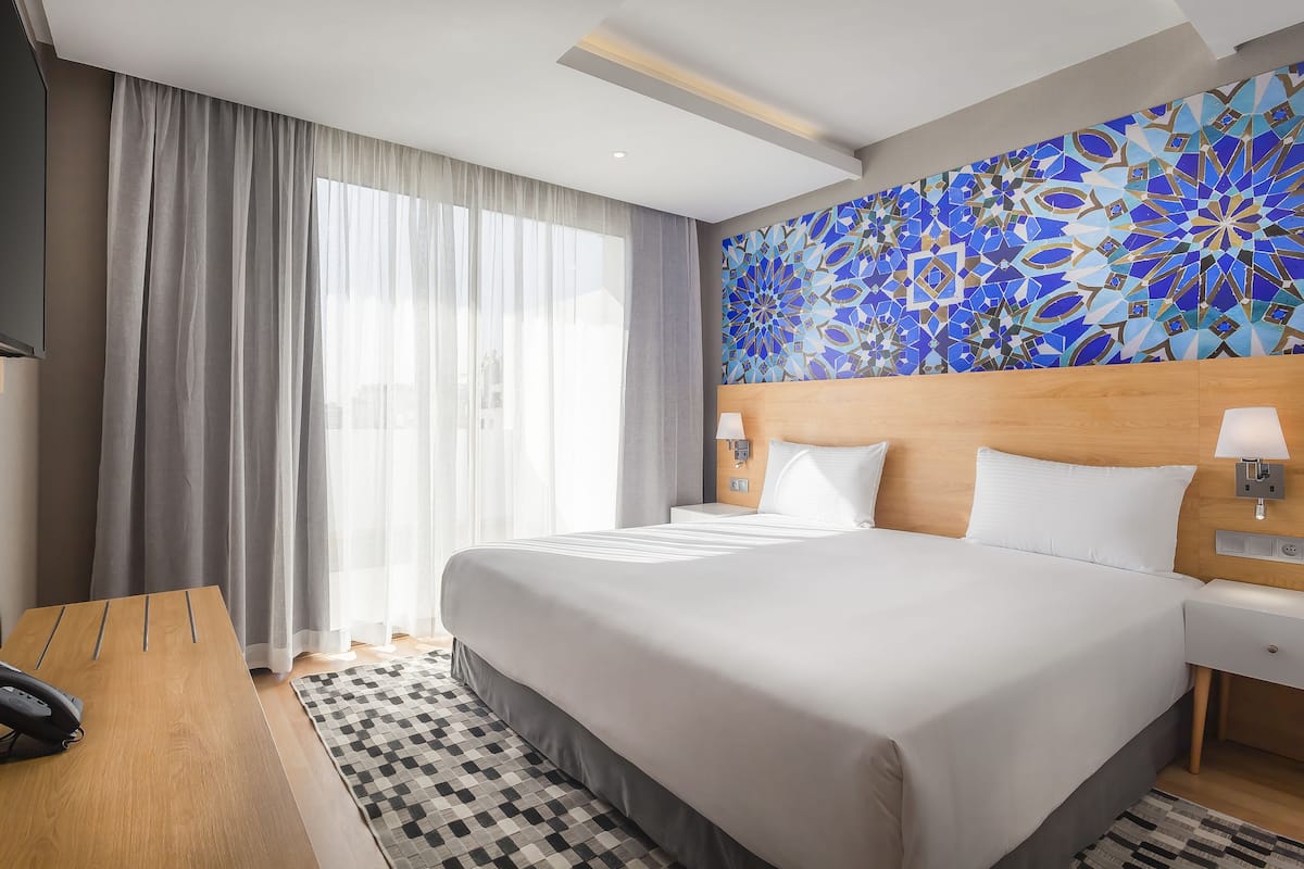 Een hotelkamer met een groot bed, een houten hoofdeinde, een geruit tapijt, een bureau met een telefoon, en een muur met een blauw-wit geometrisch patroon.