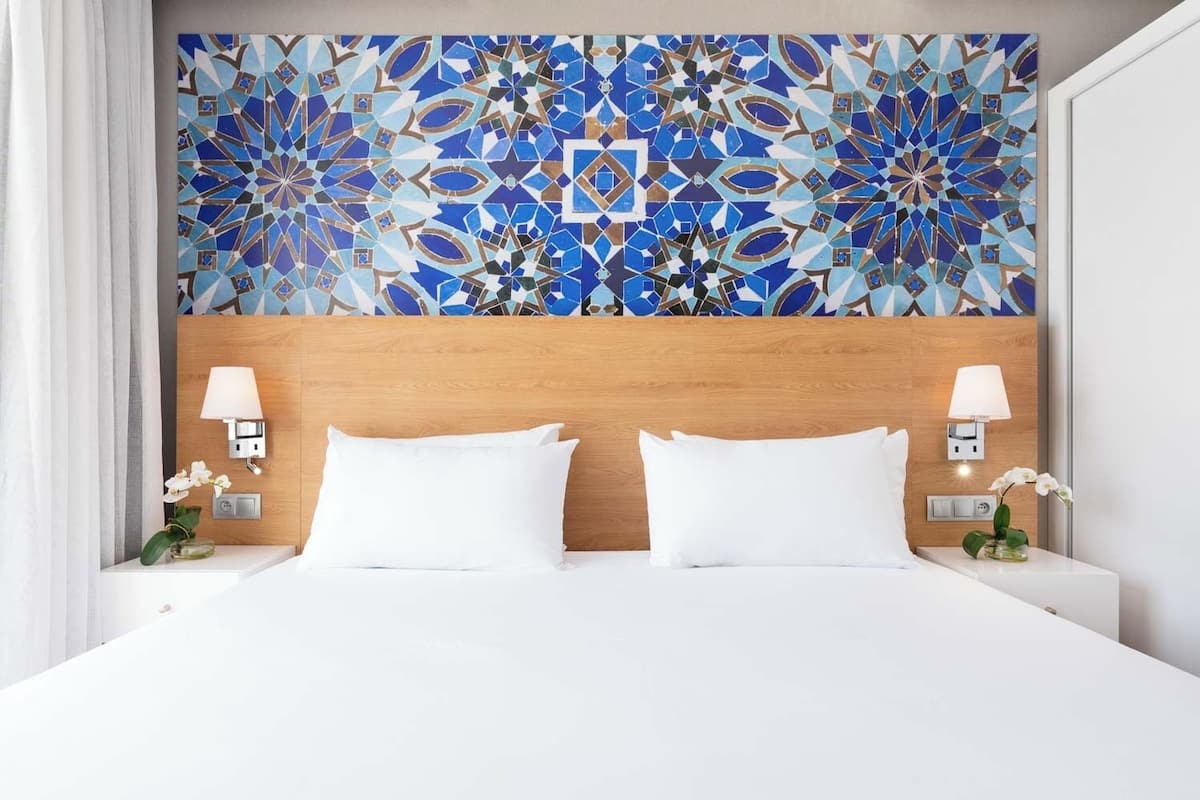 Hotelkamer met een groot bed, twee witte kussens, een houten hoofdbord en een decoratieve blauw-witte mozaïekwand.