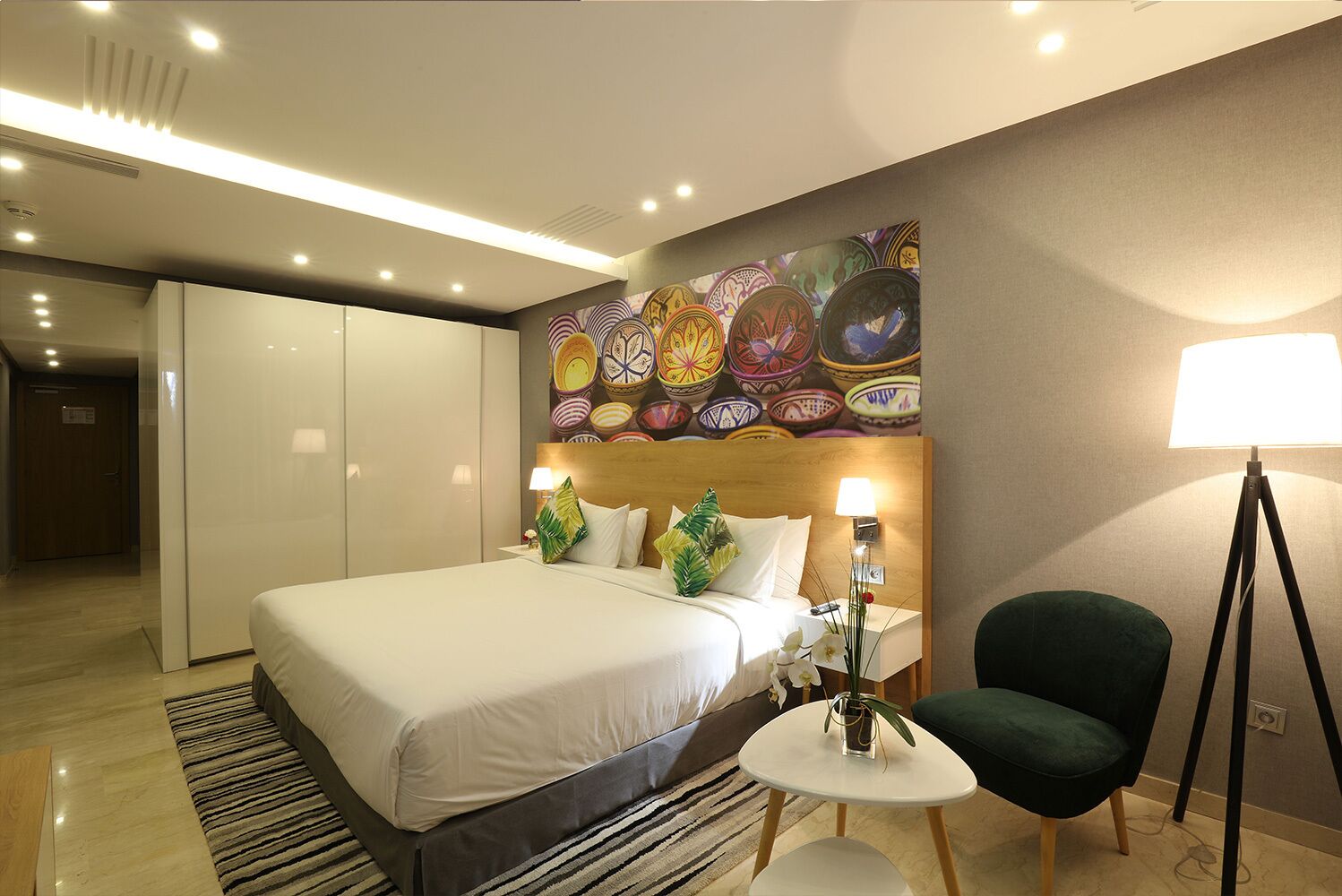 Ein Hotelzimmer mit Bett, Nachttisch, Lampe, Sessel und Wand mit farbenfrohem Wandgemälde.