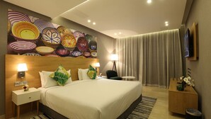 Une chambre d’hôtel moderne avec un grand lit, une tête de lit ornée d’une mosaïque colorée, un téléviseur à écran plat, une petite table d’appoint, une table de chevet, une lampe, une chaise, un vase avec des fleurs et une grande fenêtre avec des rideaux.