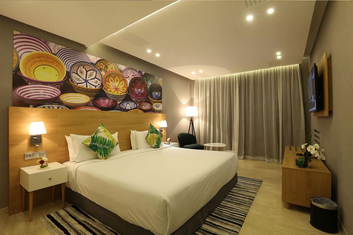 Een moderne hotelkamer met een groot bed, een hoofdbord met een kleurrijk mozaïek, een flatscreen televisie, een klein bijzettafeltje, een nachtkastje, een lamp, een stoel, een vaas met bloemen en een groot raam met gordijnen.