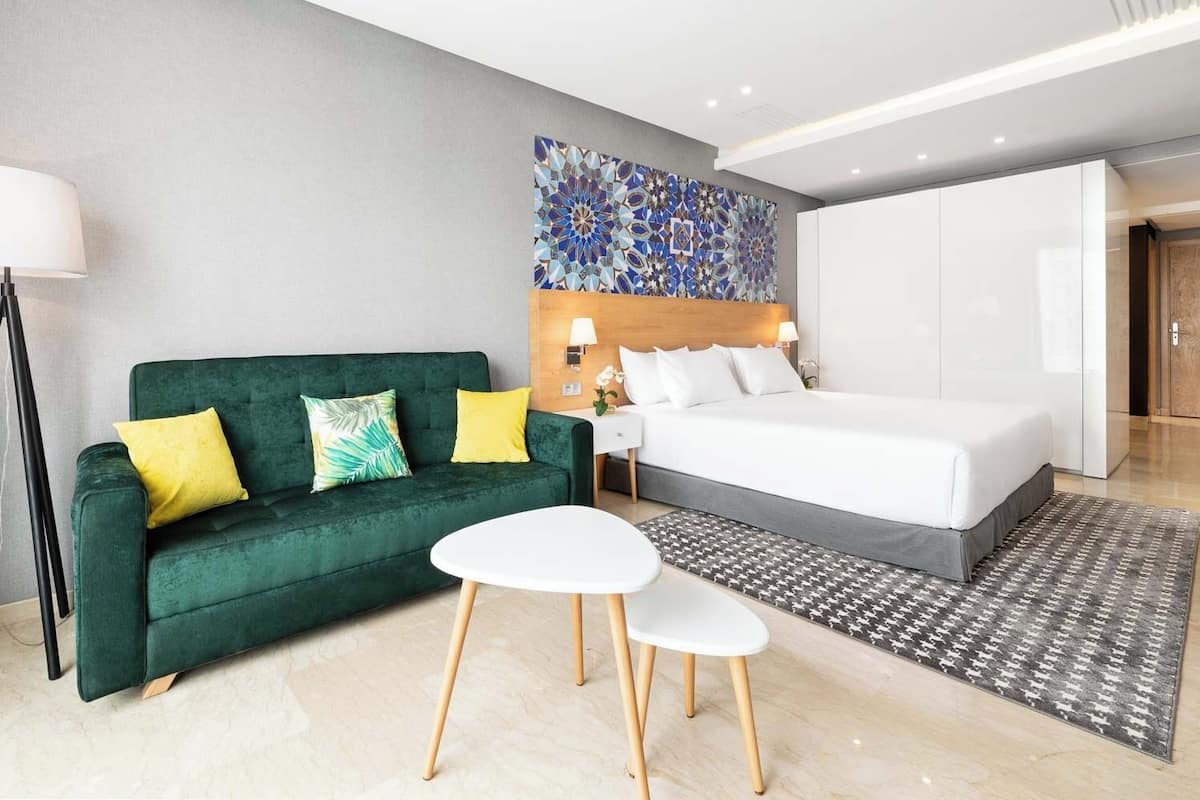 Een moderne hotelkamer met een groene bank, een wit bed, een klein wit tafeltje en een kamerbreed tapijt met een patroon.