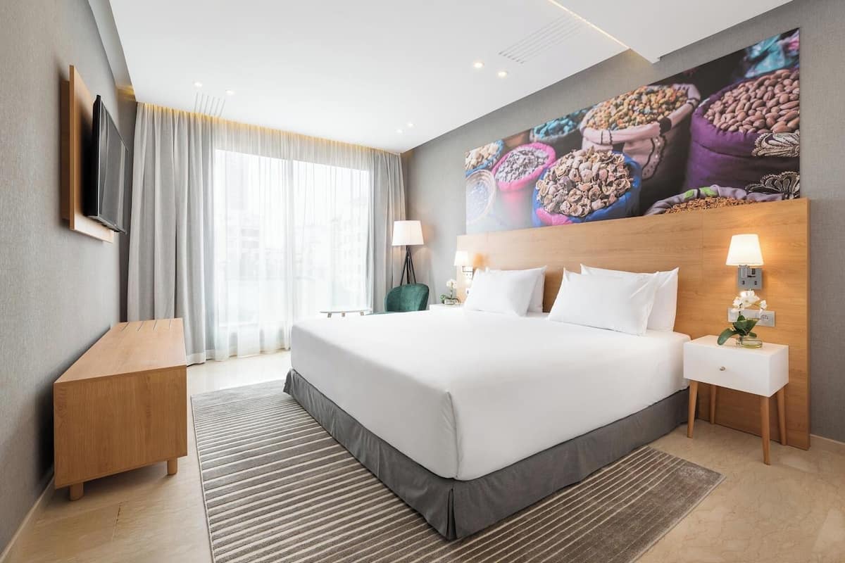 Een moderne hotelkamer met een groot bed, een flatscreen-televisie aan de wand, een houten nachtkastje en een slaapkamerkastje, plus een groot raam met dunne gordijnen.