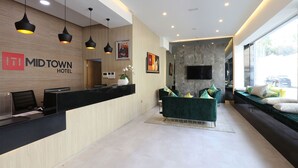 Un espace d’accueil moderne, avec un comptoir de réception en bois, des suspensions noires, un téléviseur à écran plat fixé sur un mur de marbre et un coin salon avec des canapés et des coussins verts.