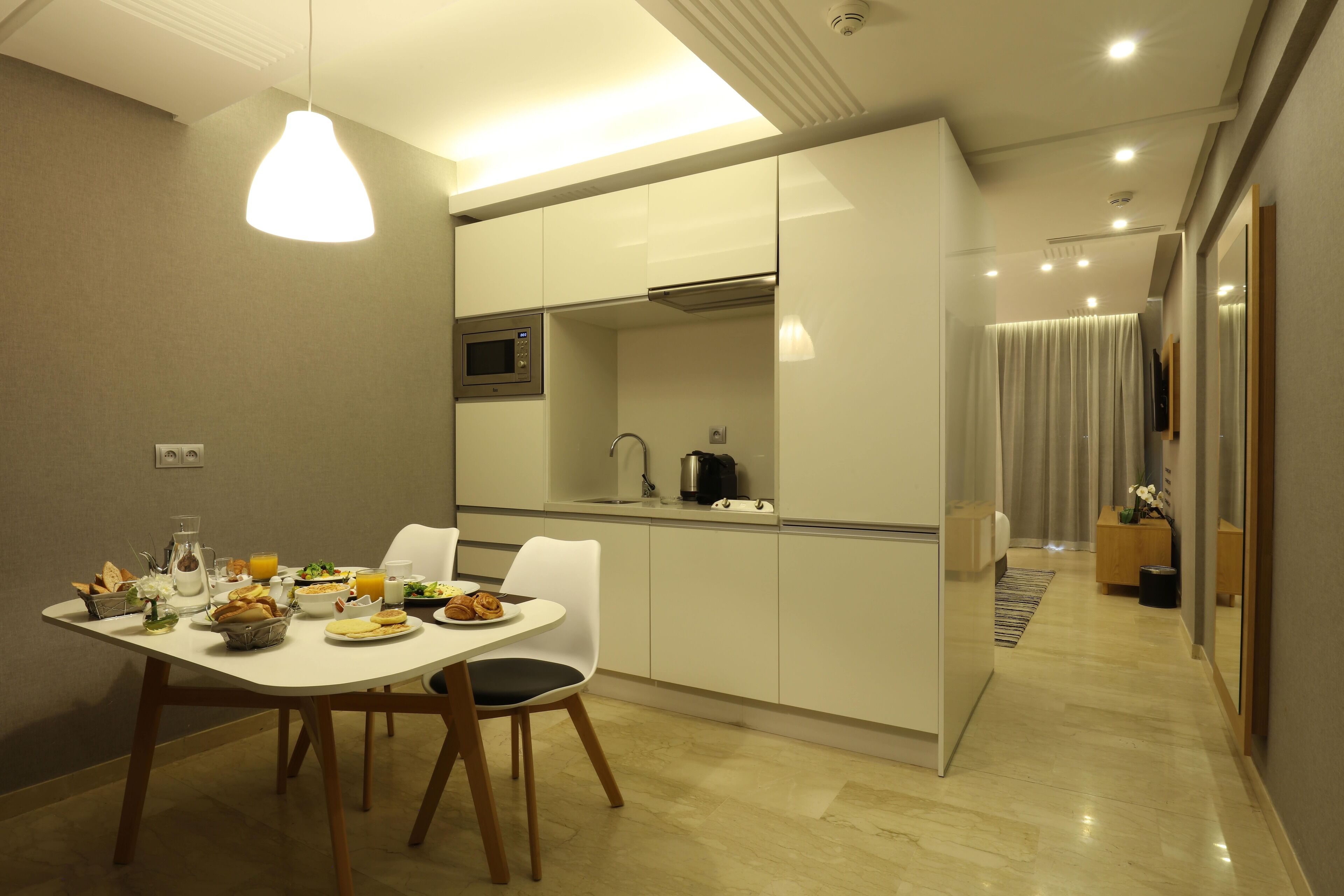 A modern kitchen with white cabinets, a microwave, and a sink. There is a dining table with chairs and a breakfast spread.