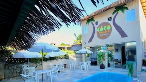 Un área de piscina con sillas de plástico blanco y sombrillas, un edificio con un mural de palmeras y un letrero que dice "Coco Beach Pousada".