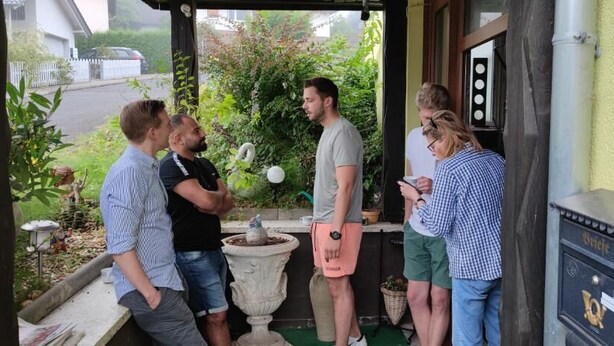 Un groupe de personnes se tient devant un bâtiment, près d’une boîte aux lettres et d’un pot de fleurs.