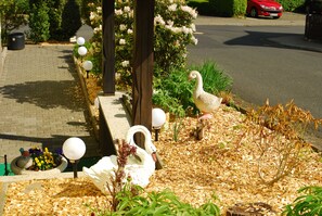 A garden with swan sculptures, a paved walkway, and a red car parked on the street.