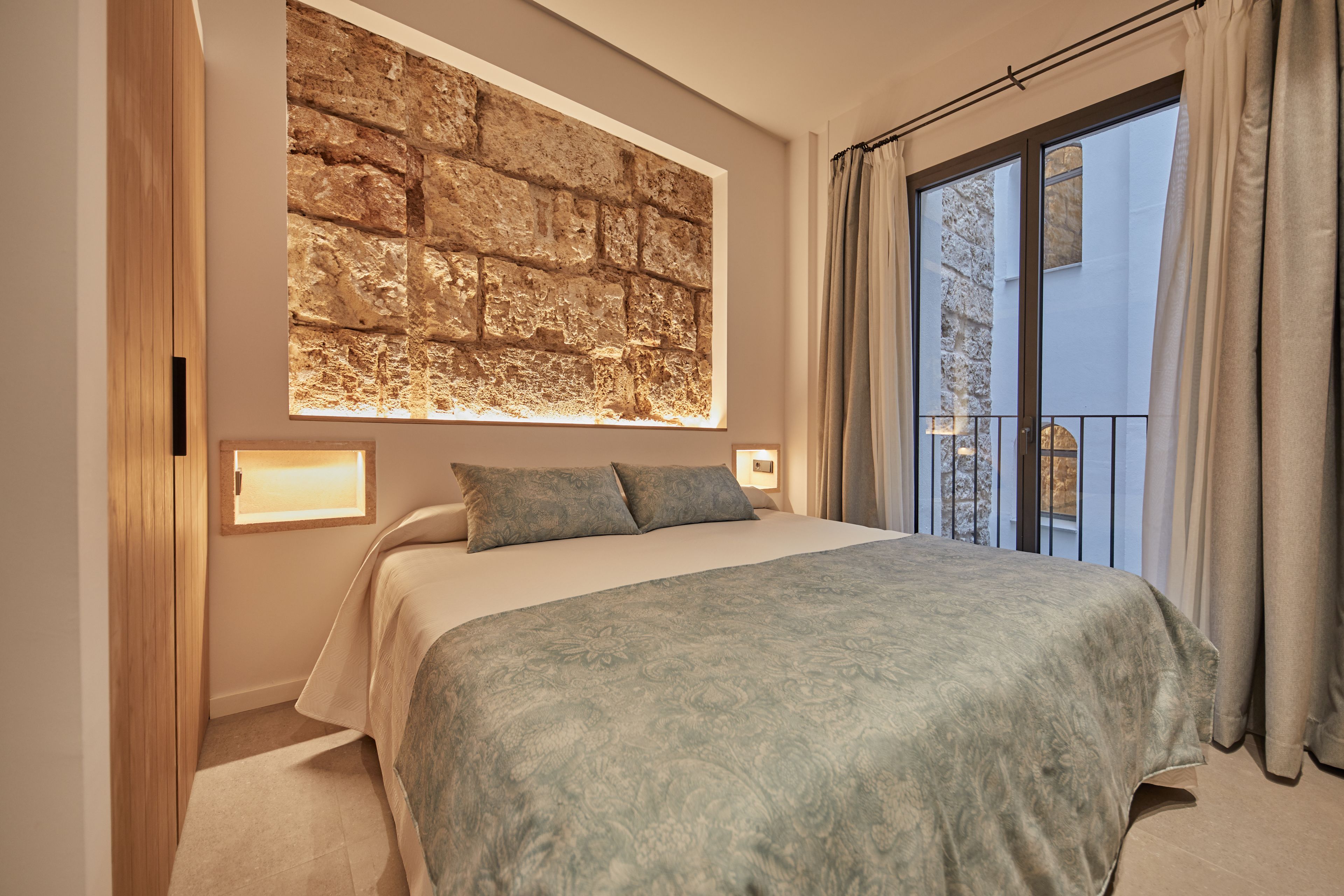 A bedroom with a large bed, a stone wall, a window with curtains, and a balcony.