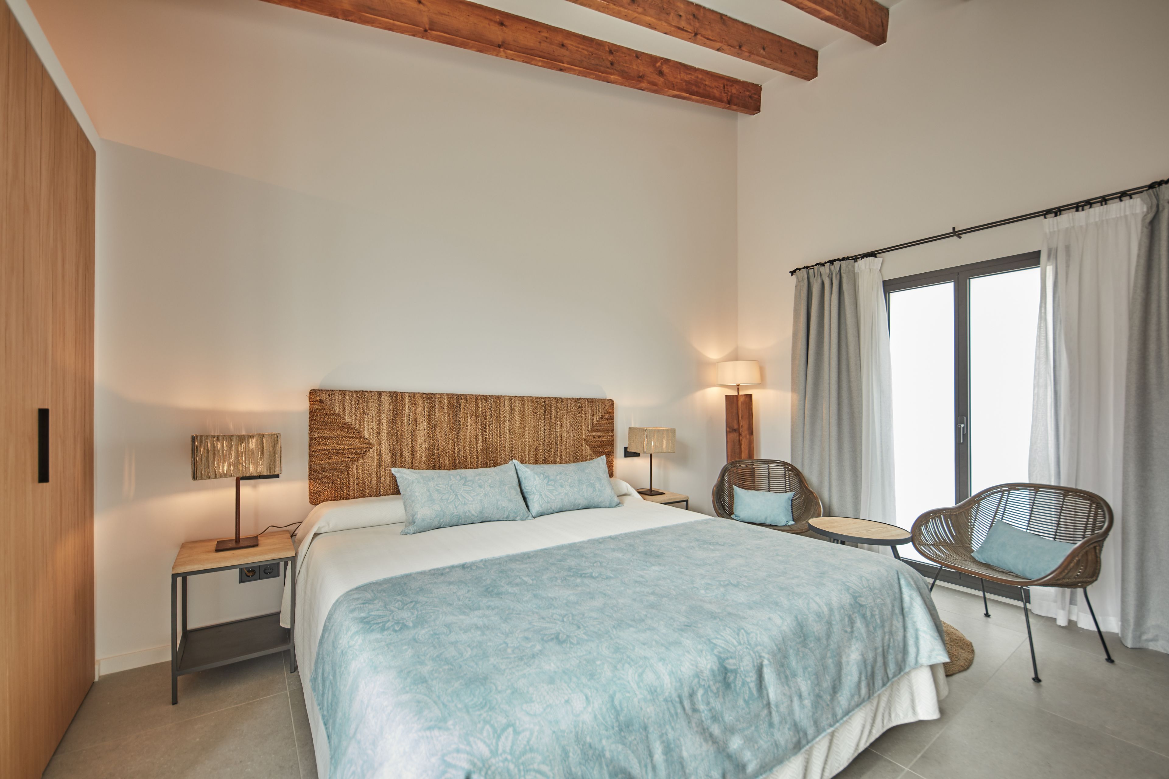 Un dormitorio con una cama grande, dos mesitas de noche con lámparas, una silla de mimbre y una ventana con cortinas.