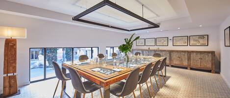 Un comedor espacioso con una gran mesa de madera, sillas modernas y una mesa con cubierta de vidrio. Se observan obras de arte enmarcadas en las paredes y un aparador de madera al fondo.