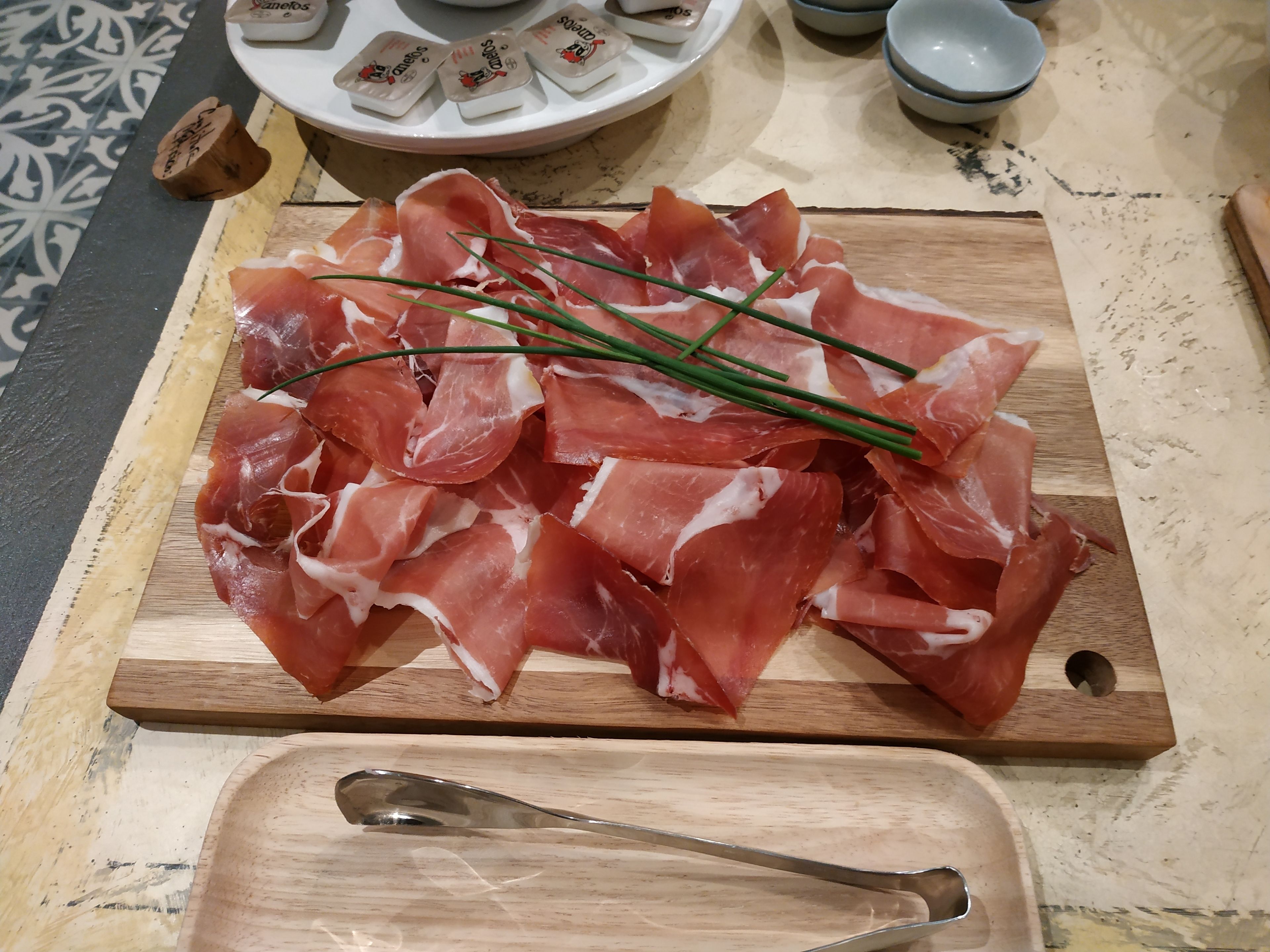 Una tabla de madera con jamón serrano en lonchas y cebollines sobre un suelo de baldosas.