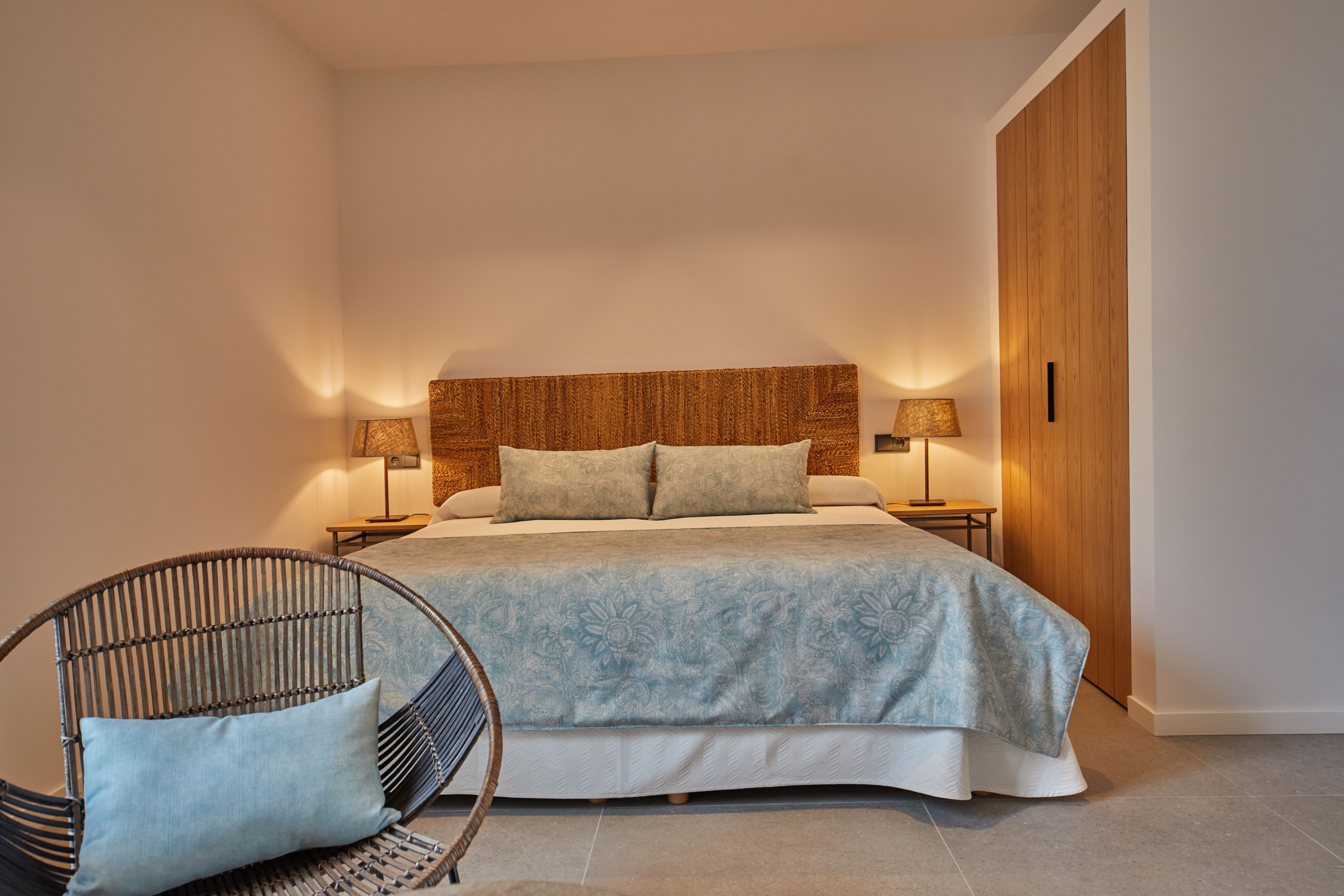 A bedroom with a large bed, two bedside tables with lamps, a wicker chair, and a wooden wardrobe.