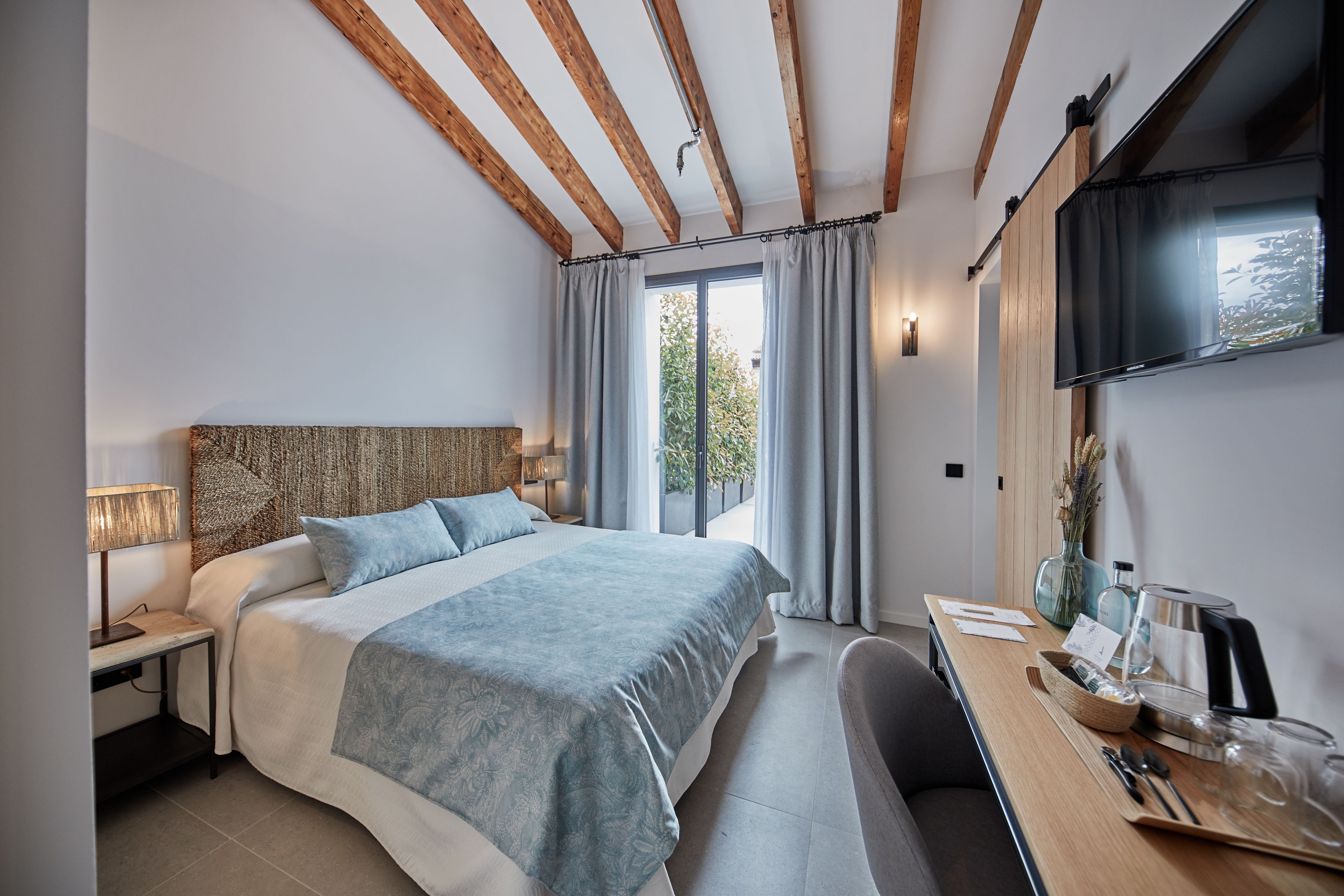 A modern bedroom with a large bed, wooden beams, a desk with a coffee maker, and a TV on the wall.