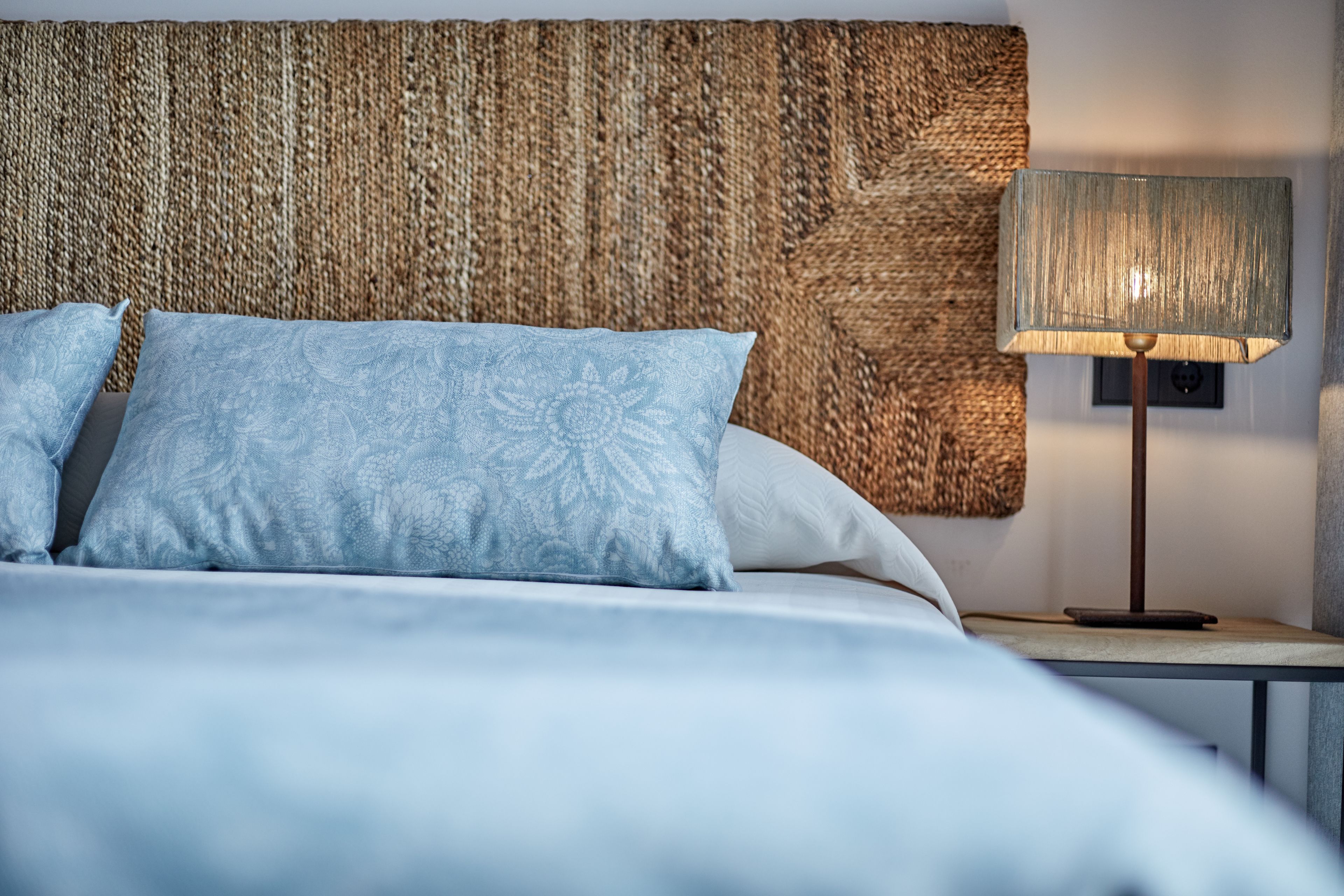 A neatly made bed with patterned pillows, a woven headboard, and a bedside lamp on a nightstand.