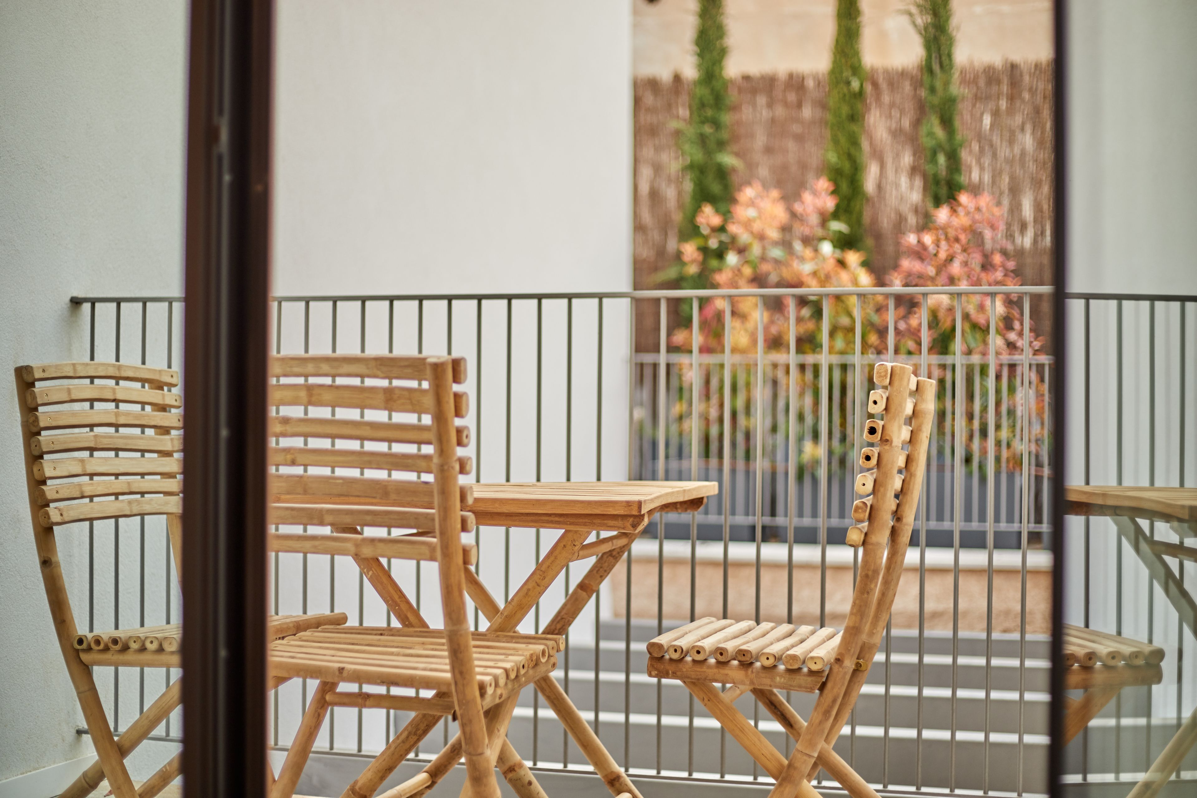 A balcony with two wooden chairs and a small table, a metal railing, and a view of a garden with flowers and trees.