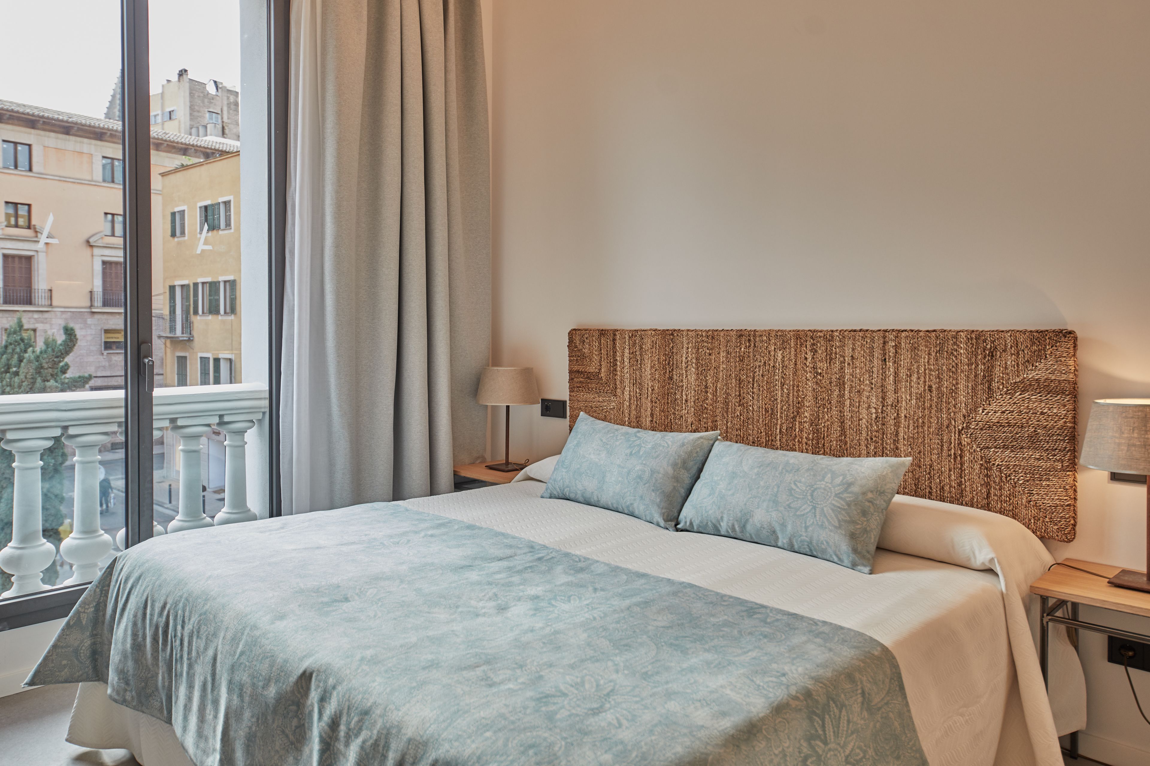 A hotel room with a large bed, a headboard with a woven texture, a window with a view of buildings, and a nightstand with a lamp.