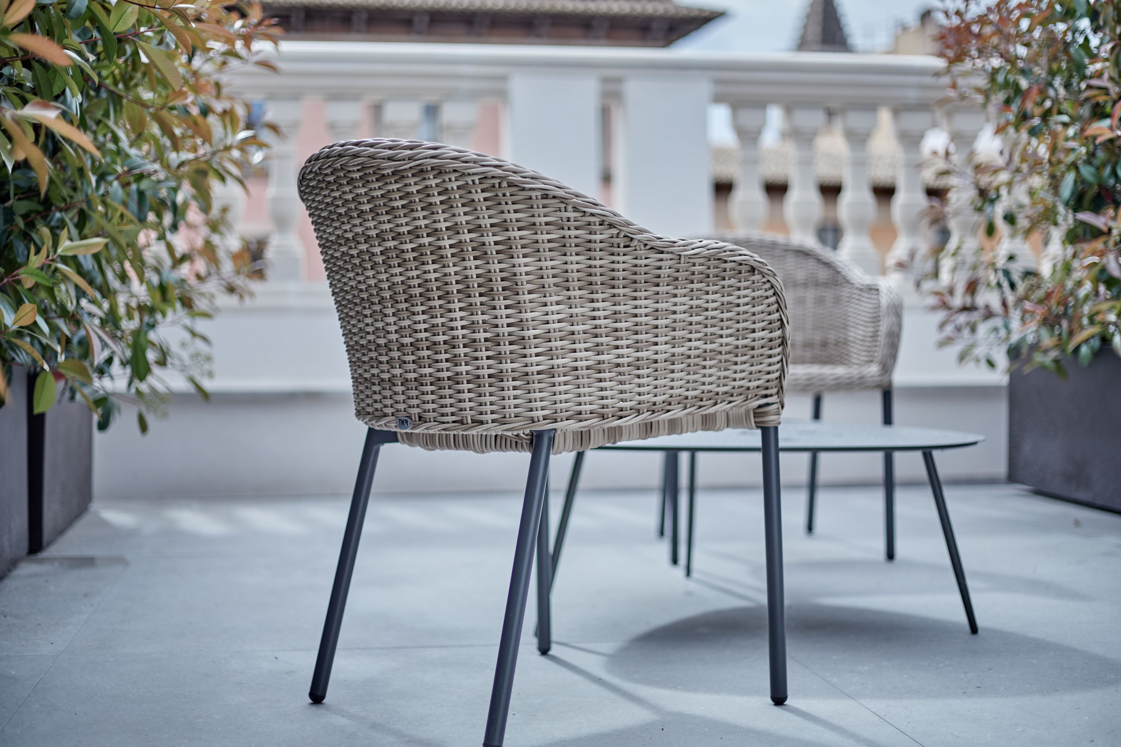 A wicker chair with metal legs on a balcony with a railing and potted plants.