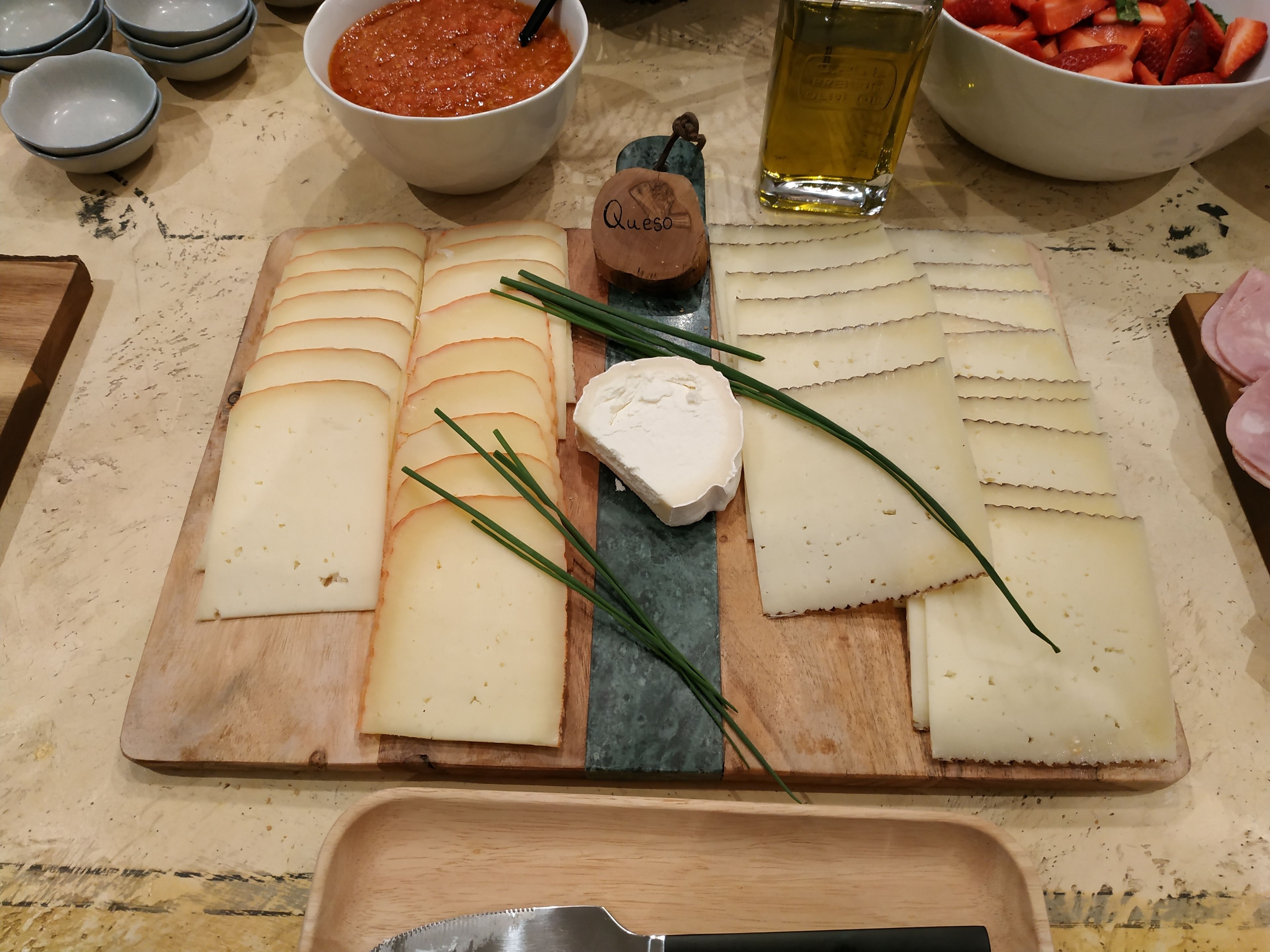 Una tabla de quesos con varios tipos de queso, cebollín y una botella de aceite de oliva sobre una tabla de madera.