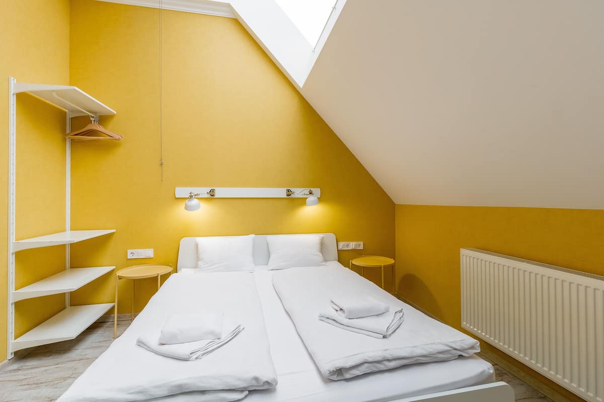 A room with a bed, white linens, a small table, a shelf, and a wall-mounted light fixture.