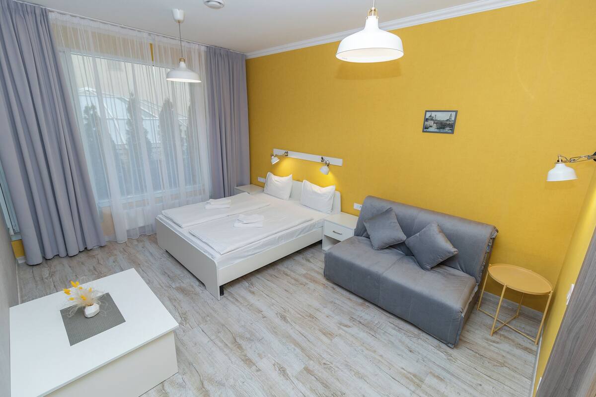 A modern hotel room with a large bed, a gray sofa, a small round table, and a white bedside table.