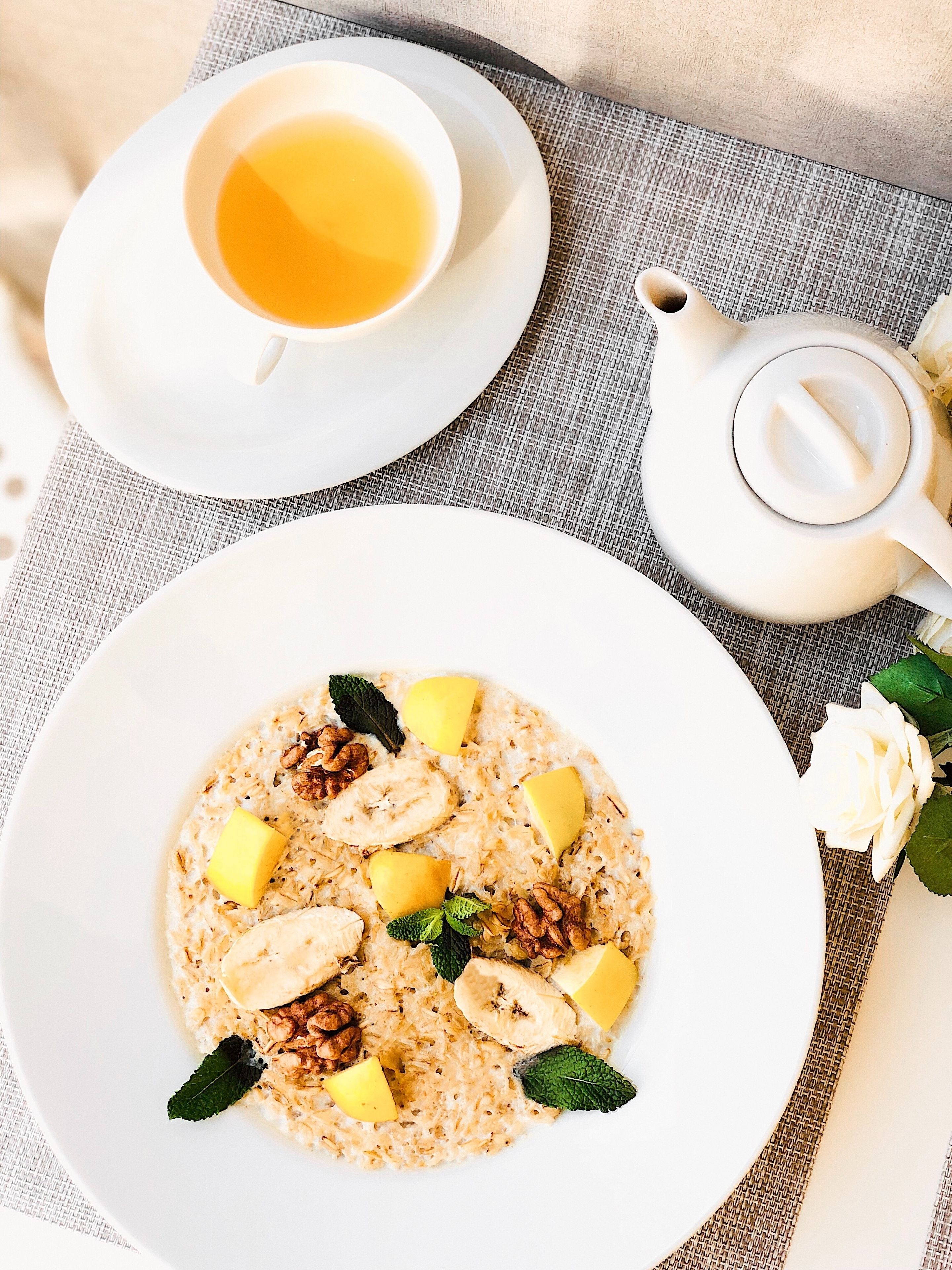 Ein Frühstückstisch mit Haferbrei, belegt mit Bananenscheiben, Walnüssen und Minzblättern, einer Tasse Tee und einer Teekanne auf einer strukturierten Tischdecke.