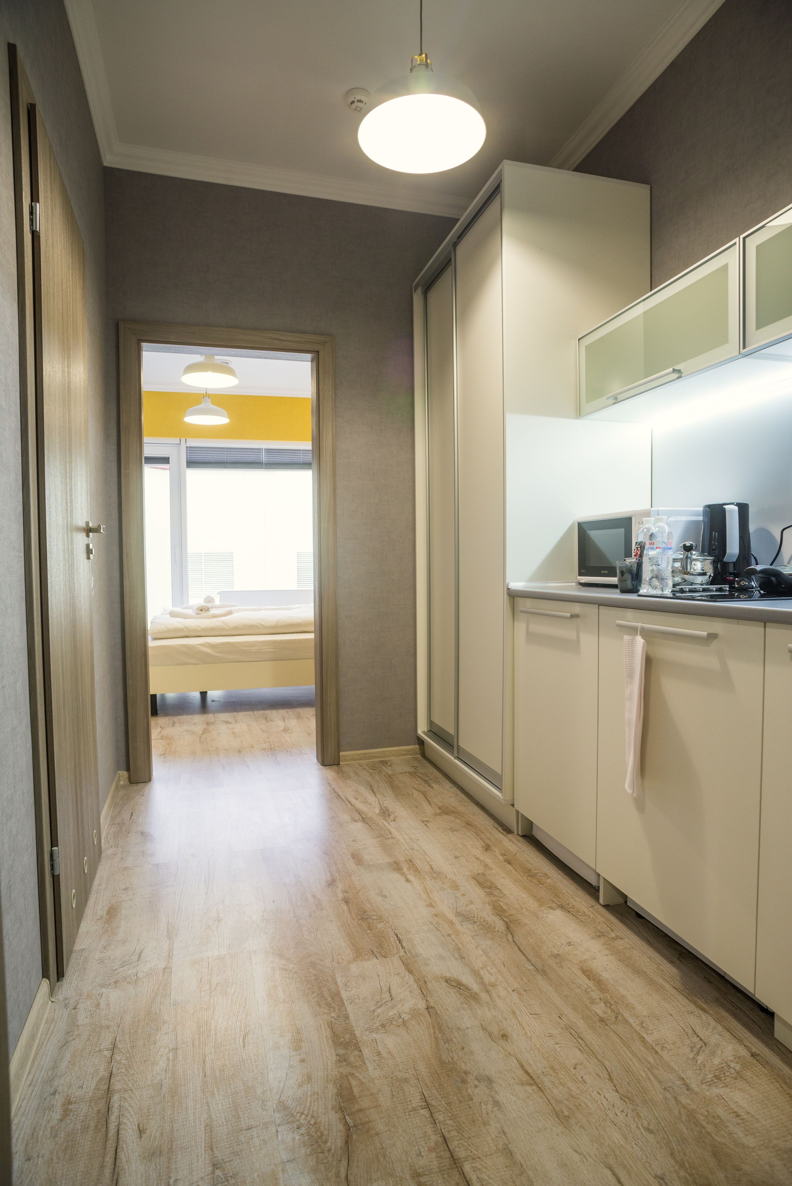 Un pasillo estrecho con suelo de madera que conduce a un dormitorio con cama y ventana. Se ve una kitchenette a la derecha, con microondas y otros electrodomésticos.