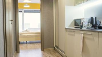 Un pasillo estrecho con suelo de madera que conduce a un dormitorio con cama y ventana. Se ve una kitchenette a la derecha, con microondas y otros electrodomésticos.
