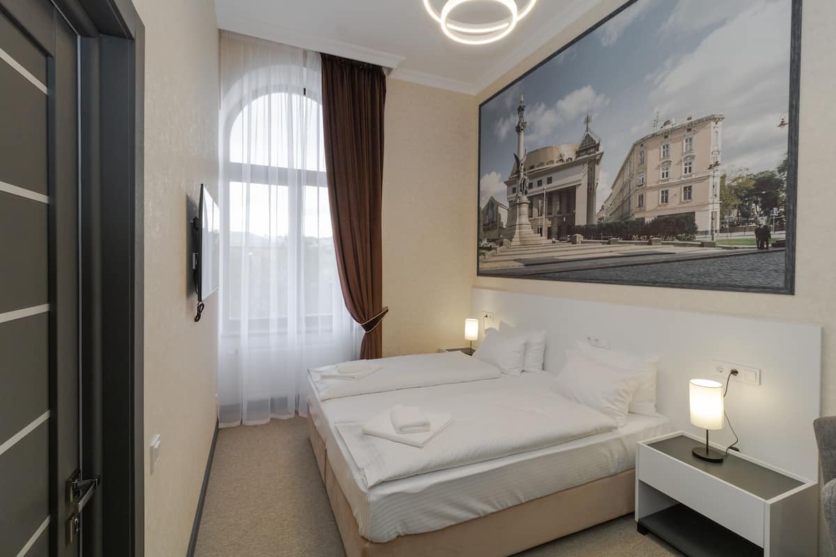 A hotel room with a large bed, a nightstand with a lamp, a painting on the wall, and a window with curtains.