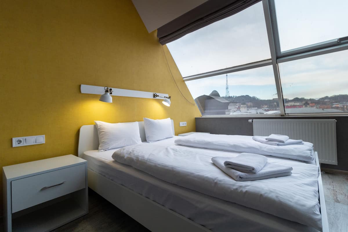 A hotel room with two beds, a nightstand, a wall-mounted lamp, and a window with a view of the cityscape.