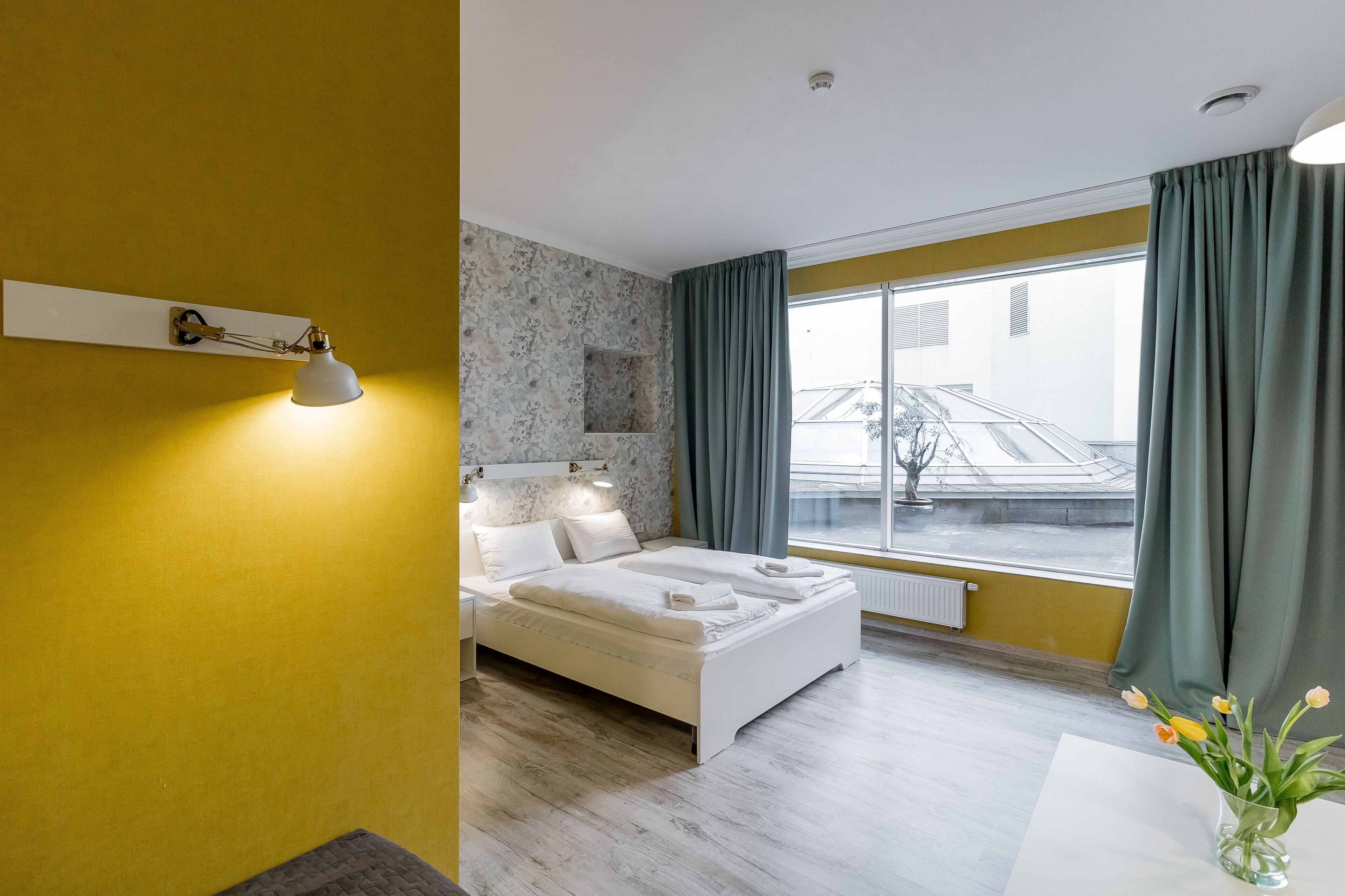 Un dormitorio moderno con cama, lámpara de noche, ventana con cortinas y vistas al exterior.