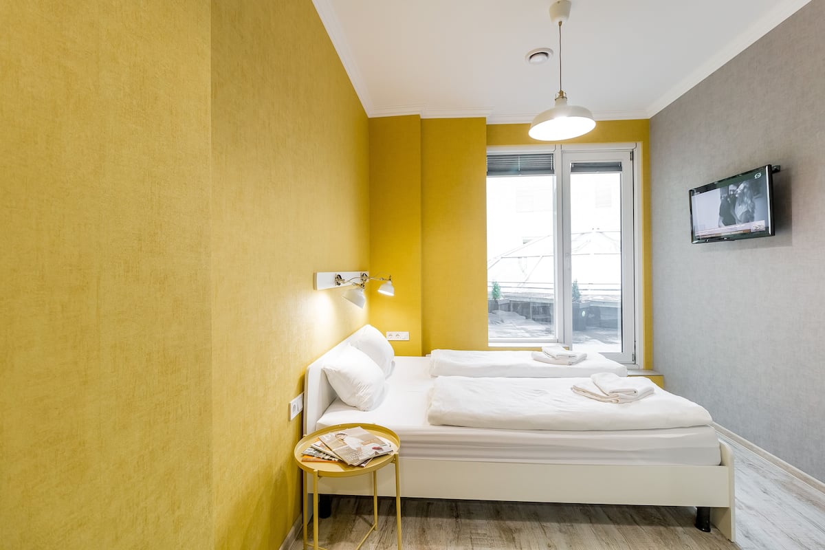 A hotel room with a bed, a small table, a window, and a TV mounted on the wall.