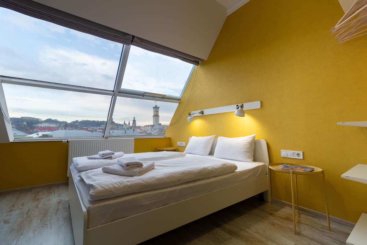 A hotel room with a large bed, a small bedside table, a window with a view of a cityscape, and a yellow accent wall.