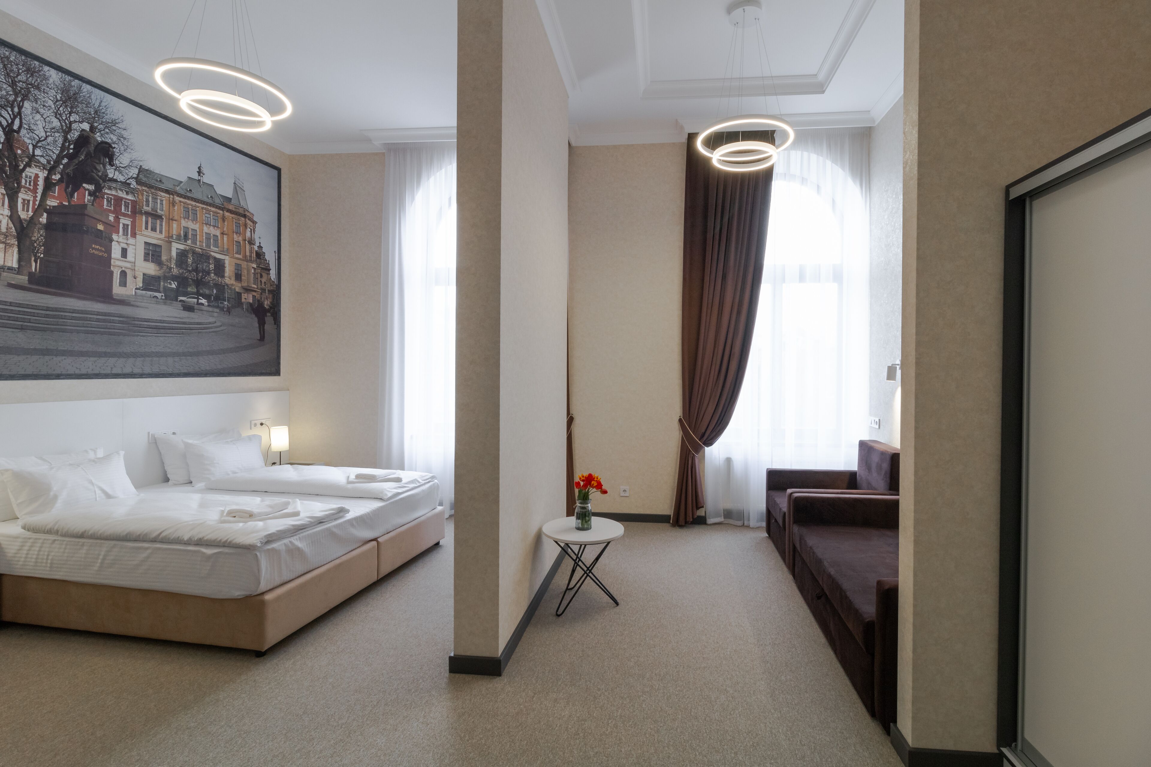 A hotel room with a bed, a small table with flowers, a sofa, and a large painting of a cityscape.