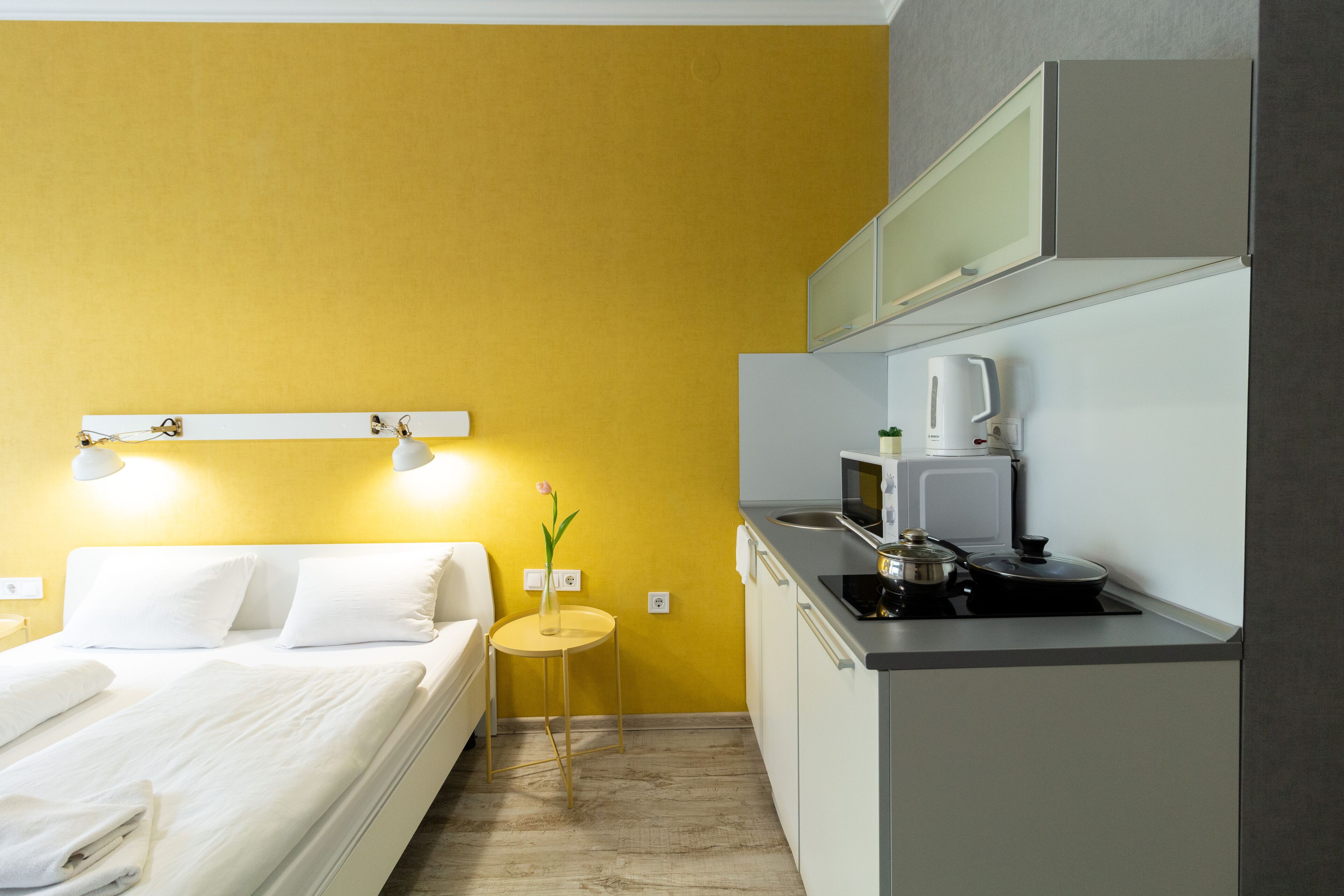 A compact hotel room with a bed, bedside table, and a small kitchenette.