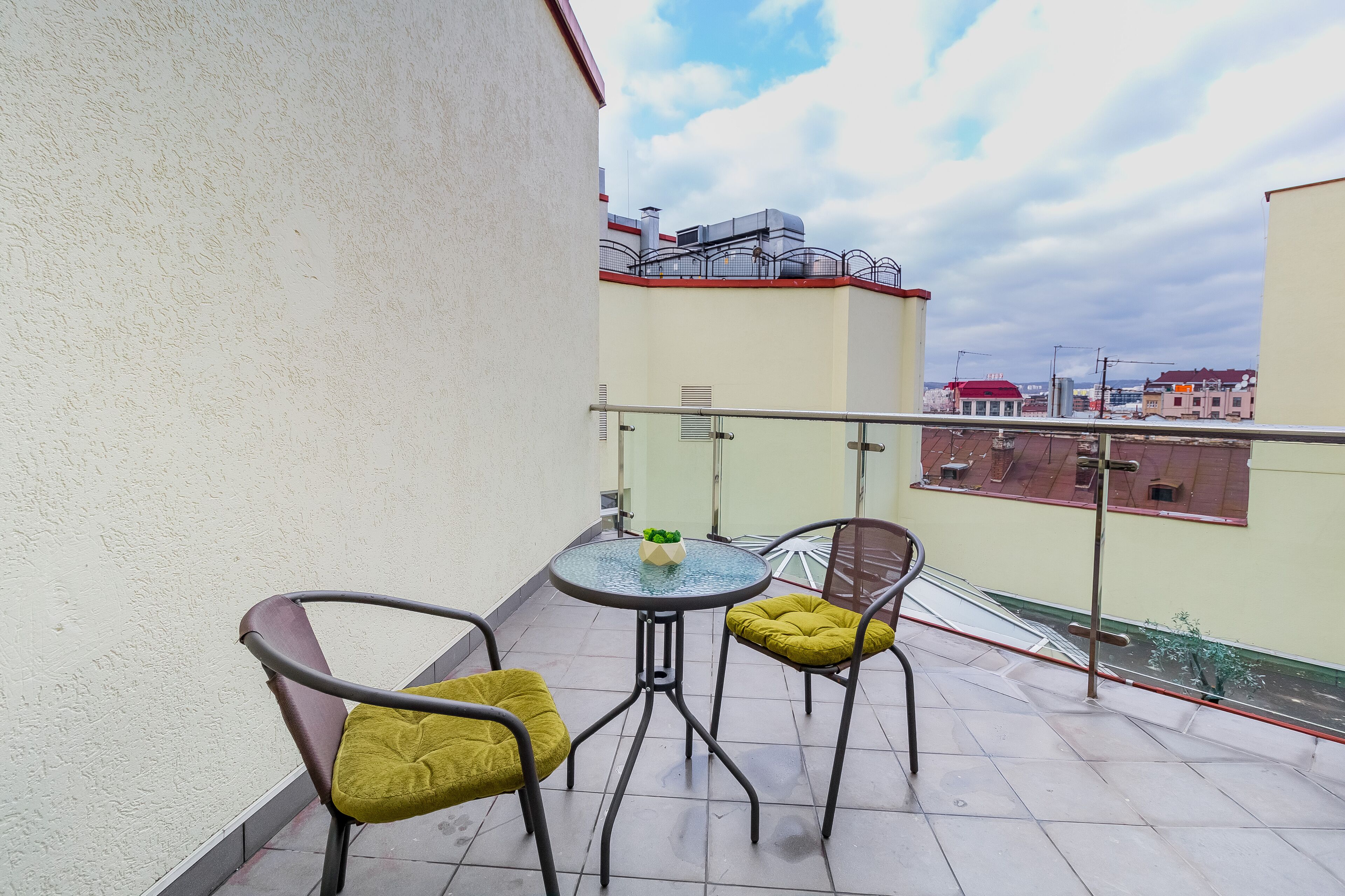 Een klein balkon met twee stoelen en een tafel, een leuning en uitzicht op gebouwen in de verte.