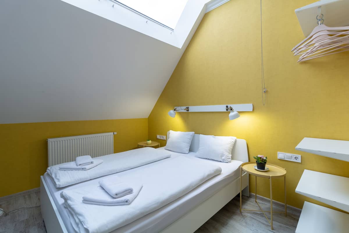 A modern bedroom with a skylight, a bed with white linens, a small bedside table with a plant, and a wall-mounted shelf.