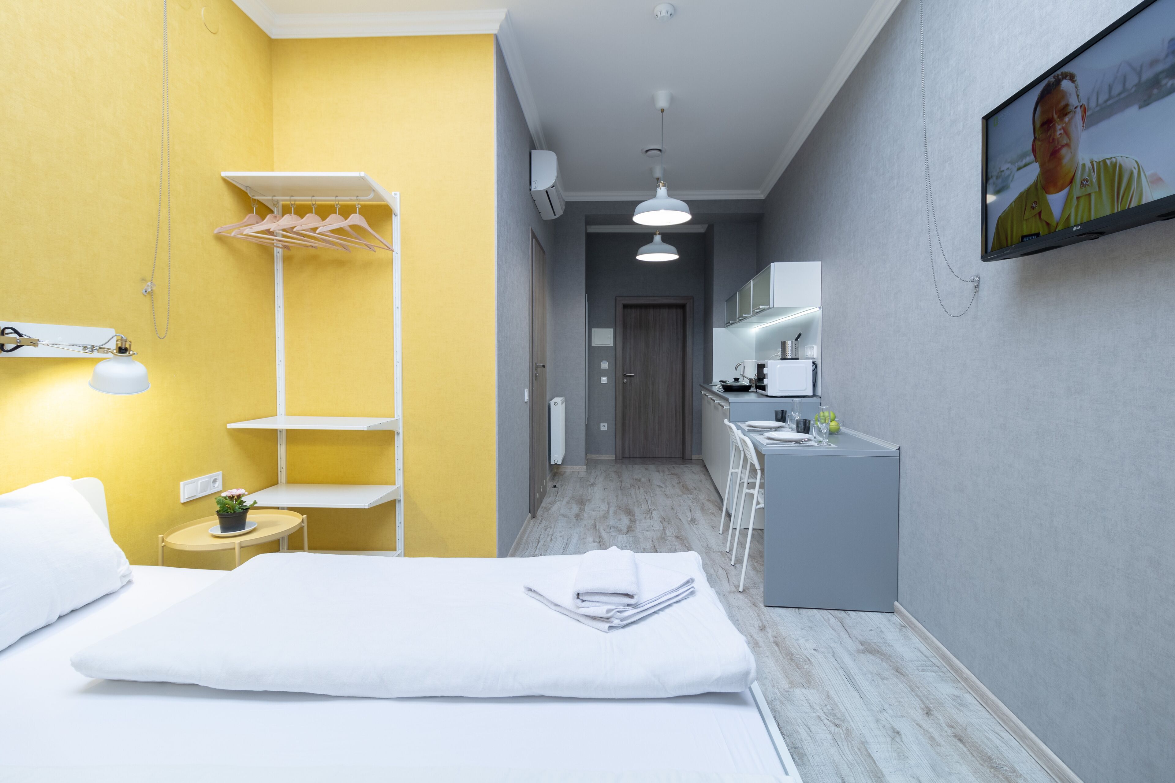 Habitación de hotel con una cama, una mesita de noche con lámpara, una planta pequeña, una toalla, un perchero y un espejo, además de un televisor en la pared.