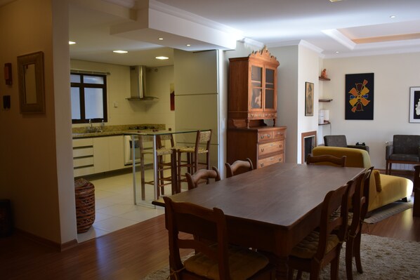 A spacious dining area with a wooden table and chairs, a kitchen in the background, and a living room with a sofa and a cabinet.