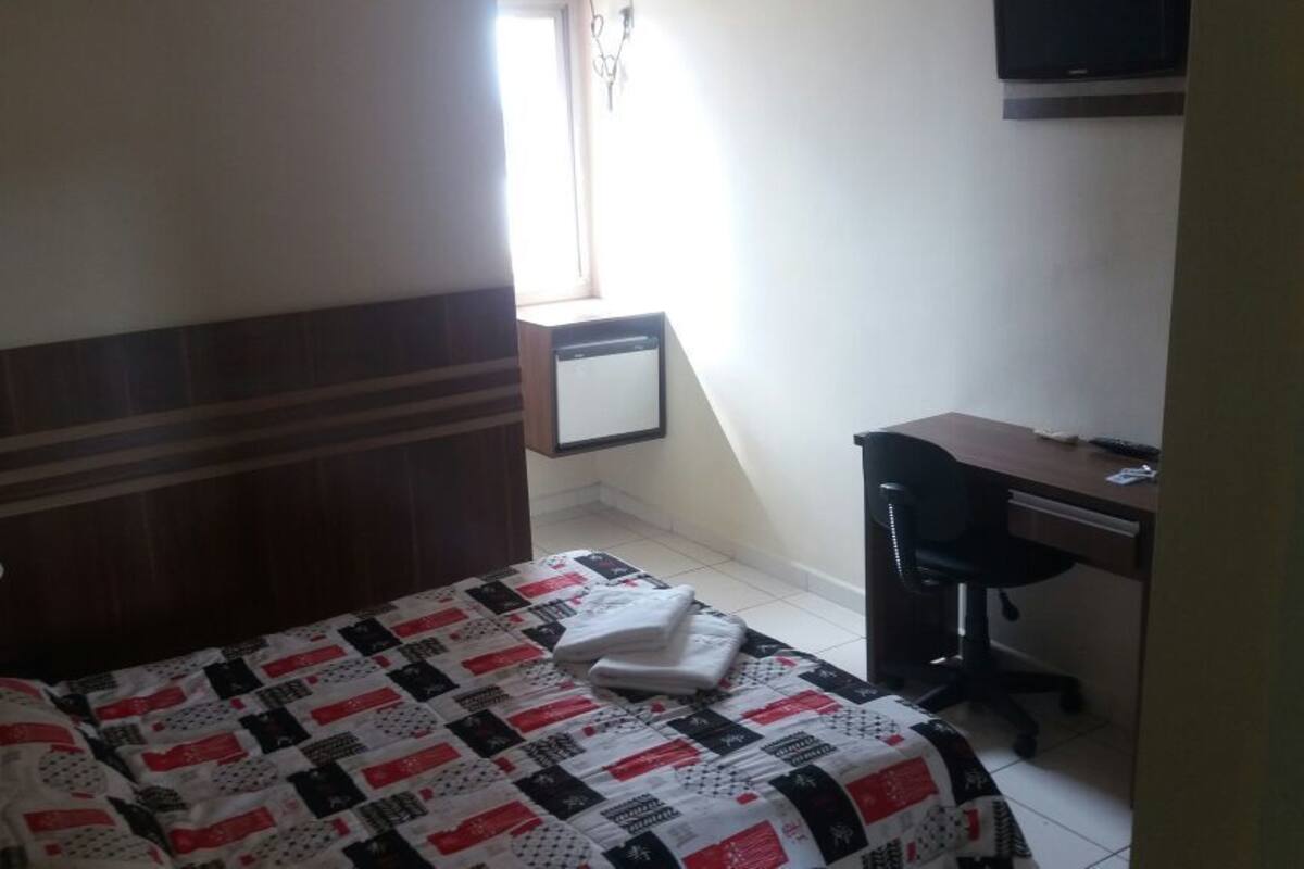 A hotel room with a bed, a desk, a chair, a lamp, and a TV.