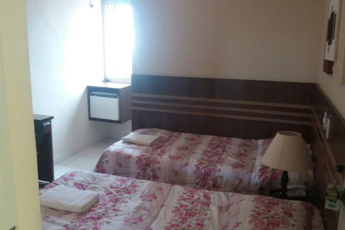 A hotel room with two beds, a nightstand, a lamp, and a window with curtains.