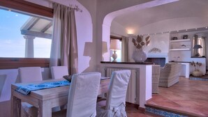 A cozy dining area with a wooden table, white chairs, and a view of the sea through a window with curtains. - Luxury Villa's Valle dell'Erica (Santa Teresa di Gallura)