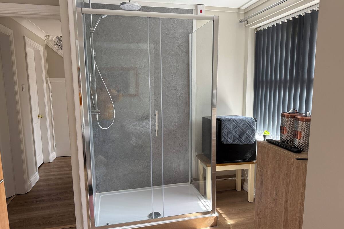 A glass-enclosed shower area with a showerhead and control panel, adjacent to a wooden chair and a small table with a keyboard and a plant.