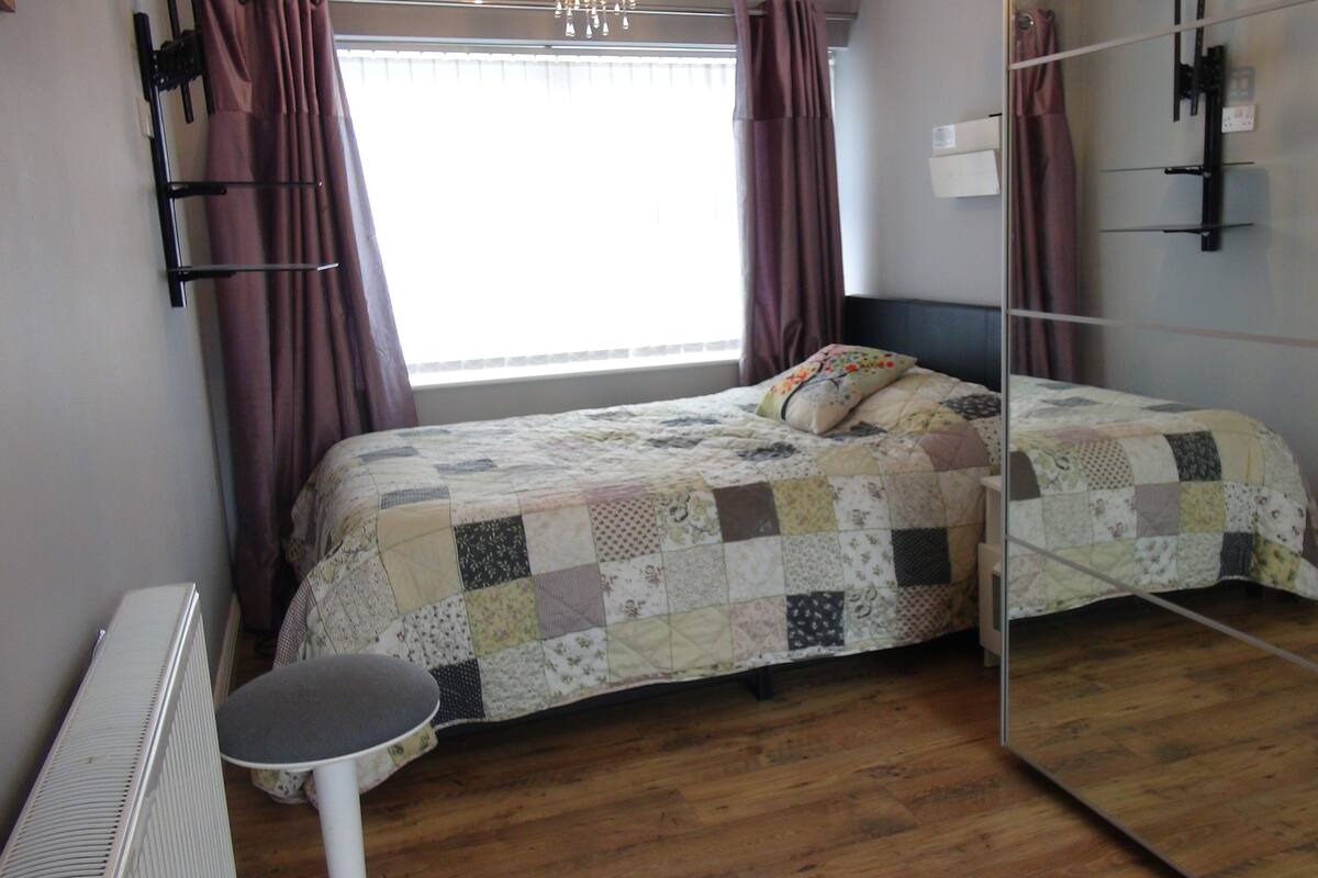 A bedroom with a wooden floor, a bed with a patchwork quilt, a small round table, and a mirror on the wall.