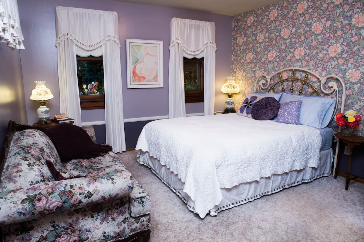 A bedroom with a floral-patterned sofa, a white bed with a decorative headboard, and a floral wallpaper.
