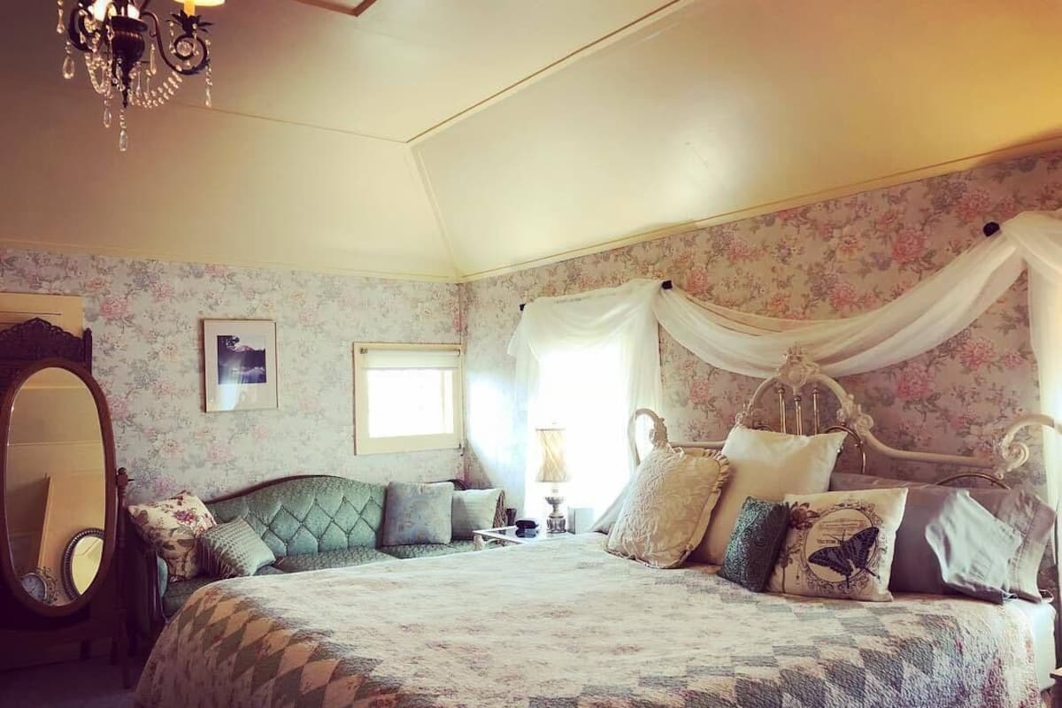 A bedroom with a large bed, floral wallpaper, a chandelier, and a mirror.