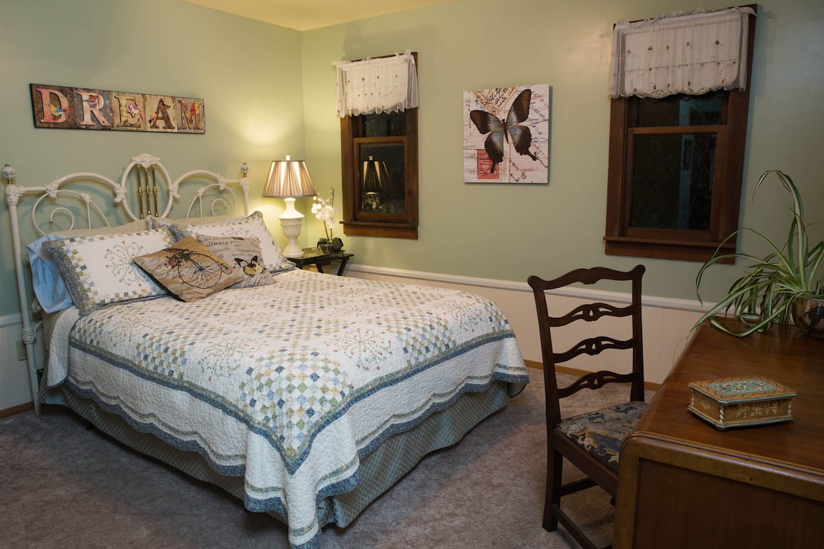 A bedroom with a bed, a nightstand, a lamp, a chair, and a table with a book.