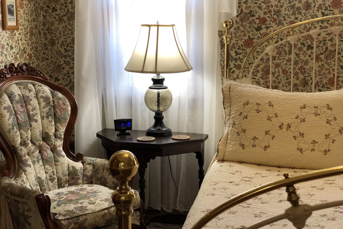 A bedroom with a floral-patterned bed, a wooden chair, a table with a lamp, and a nightstand with a clock.